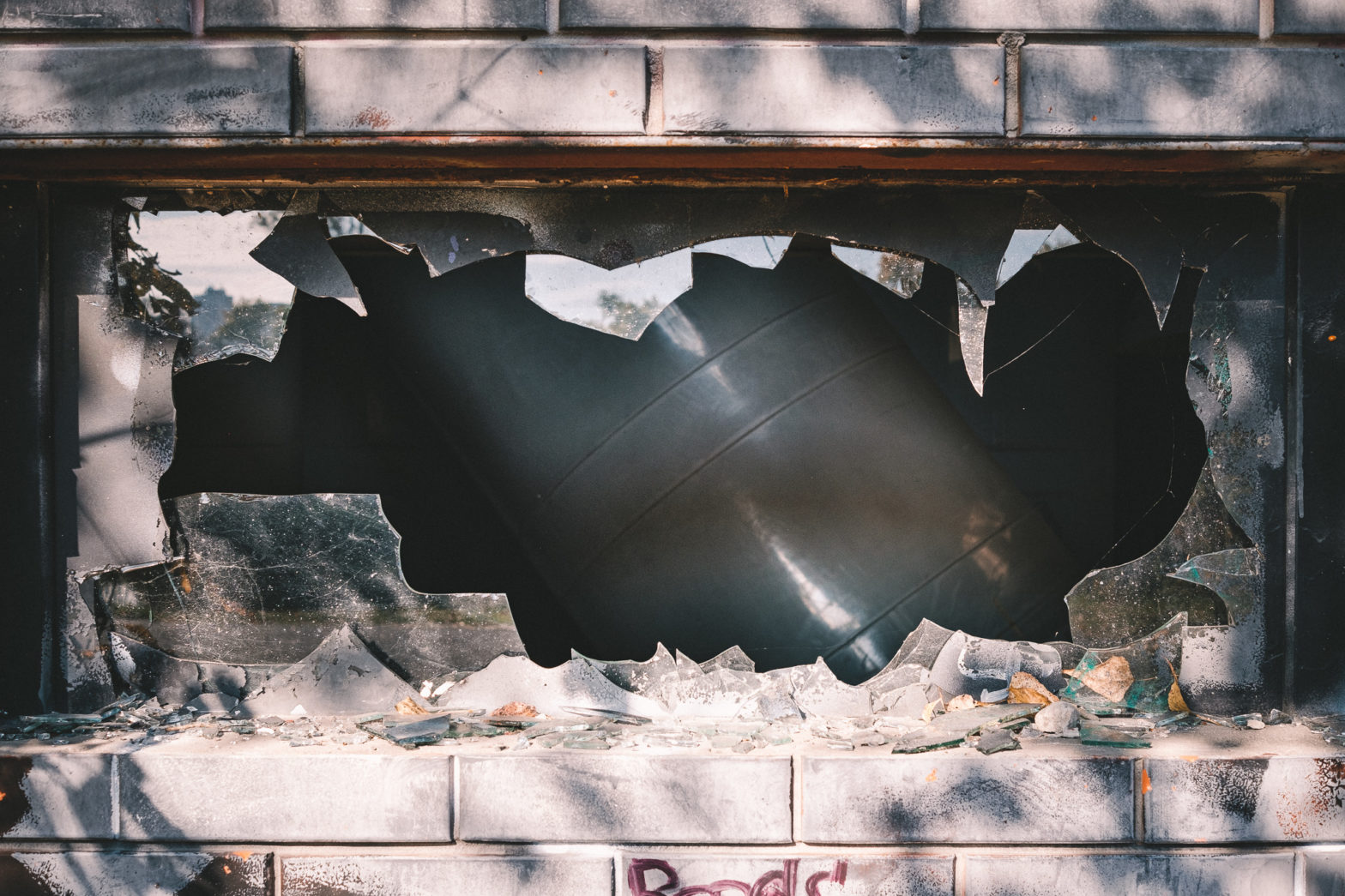A large pipe running diagonally across the room visible through a broken window with jagged shards of glass framing the view