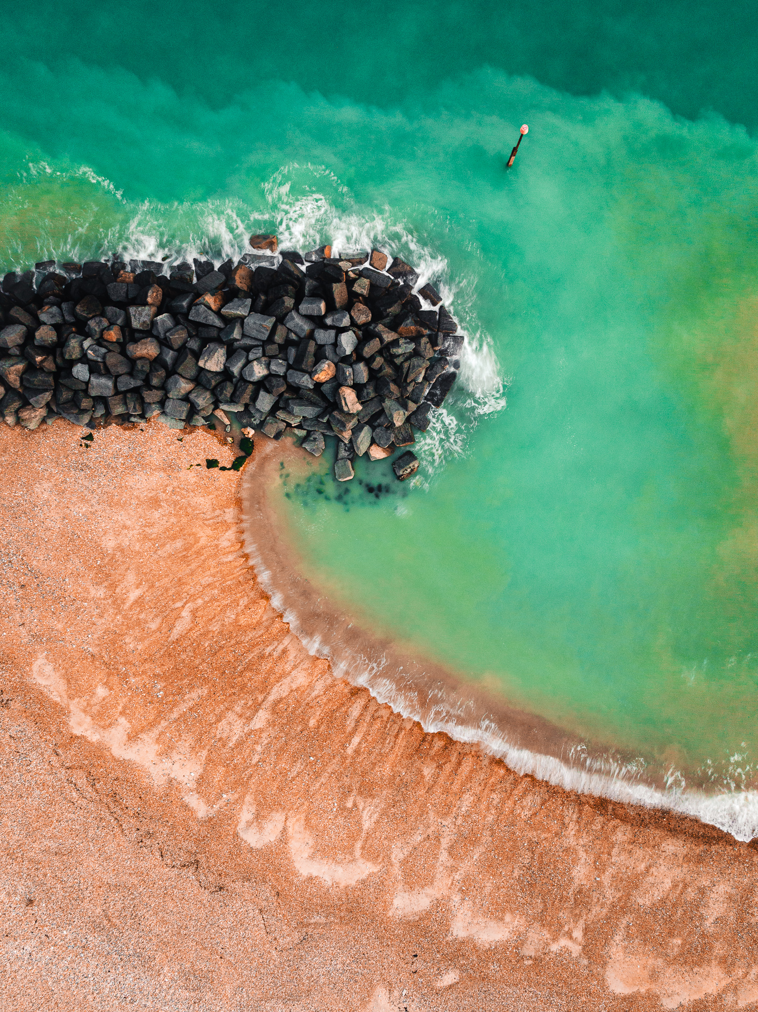 Top down view of a teal green water washing over a wave breaker made of large dark rocks with a orange pebble beach behind it