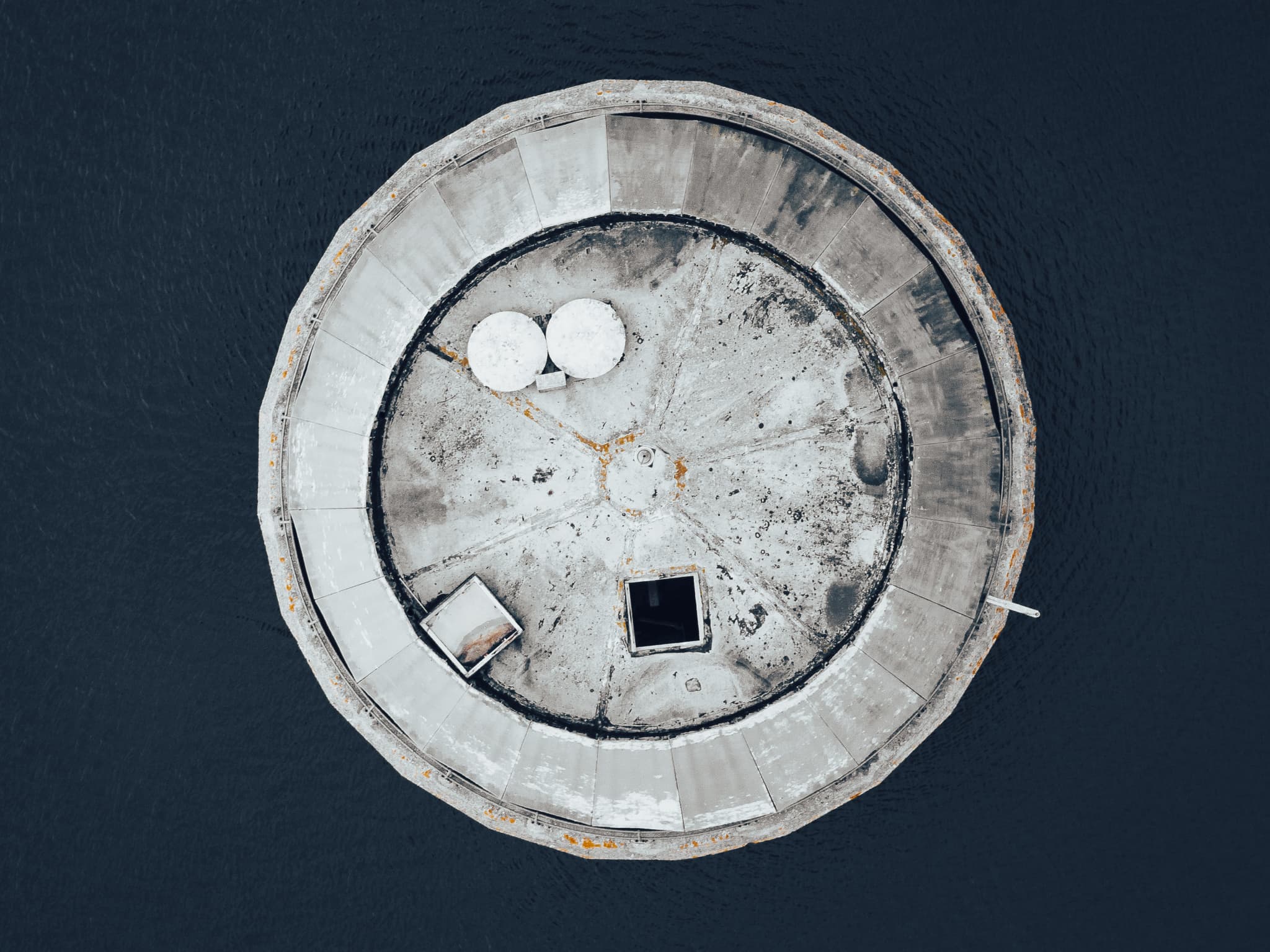 An aerial closeup view of a cement structure in the middle of a water reservoir in deep blue desecrated colours
